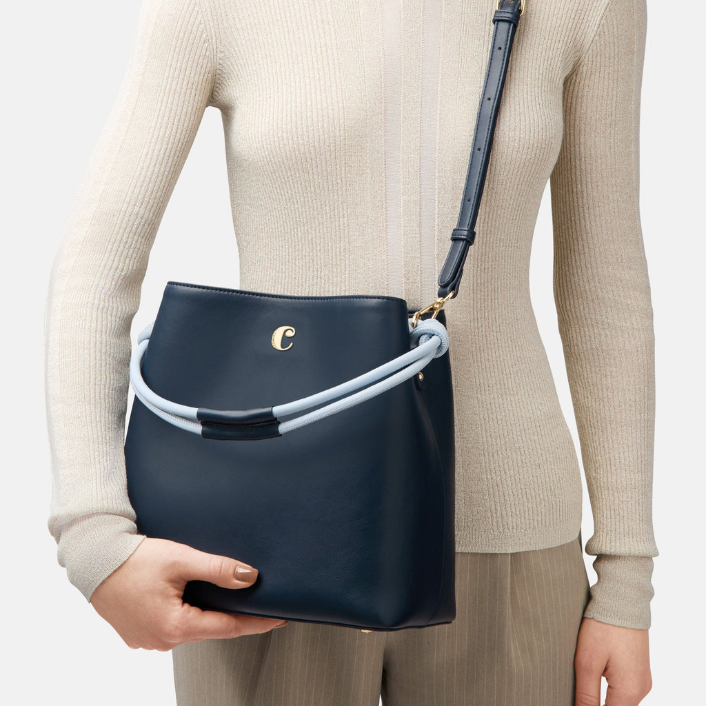 Person holding a navy blue handbag with Cacharel metal logo against a neutral background